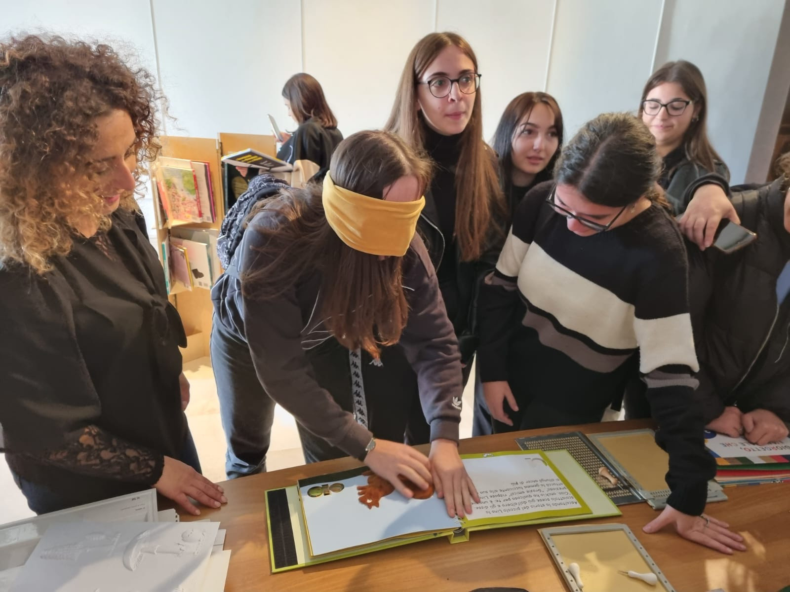 Un gruppo di ragazzine è riunito attorno a una scrivania mentre esplora un libro tattile. Una delle bambine, bendata con una fascia gialla, prova a leggere con il tatto, mentre le compagne osservano curiose.