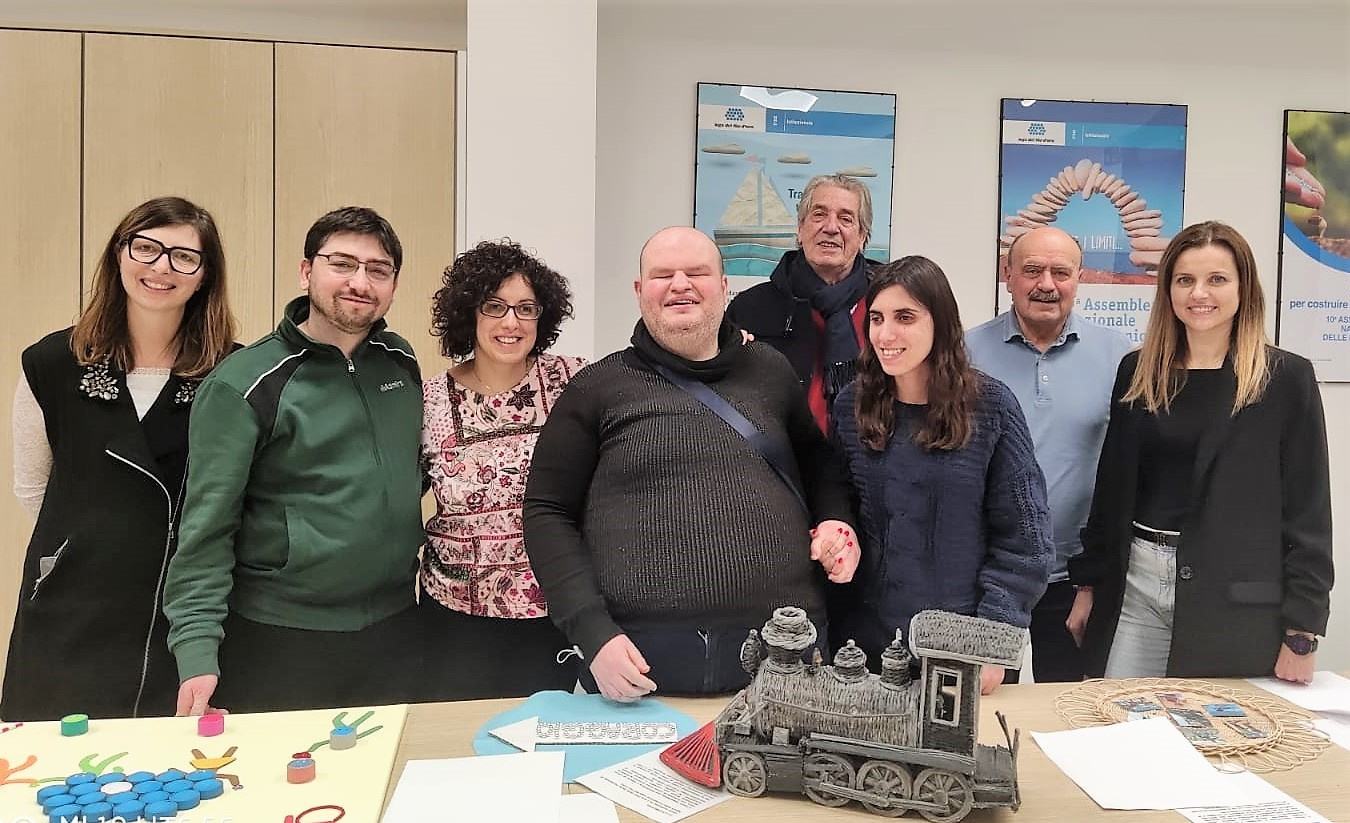 Foto di gruppo con utenti e operatori della Lega del Filo d’Oro, sorridenti mentre mostrano un’opera tattile: una locomotiva realizzata con carta arrotolata in piccoli cordoncini. Il corpo è dipinto di nero, la testata di rosso. 