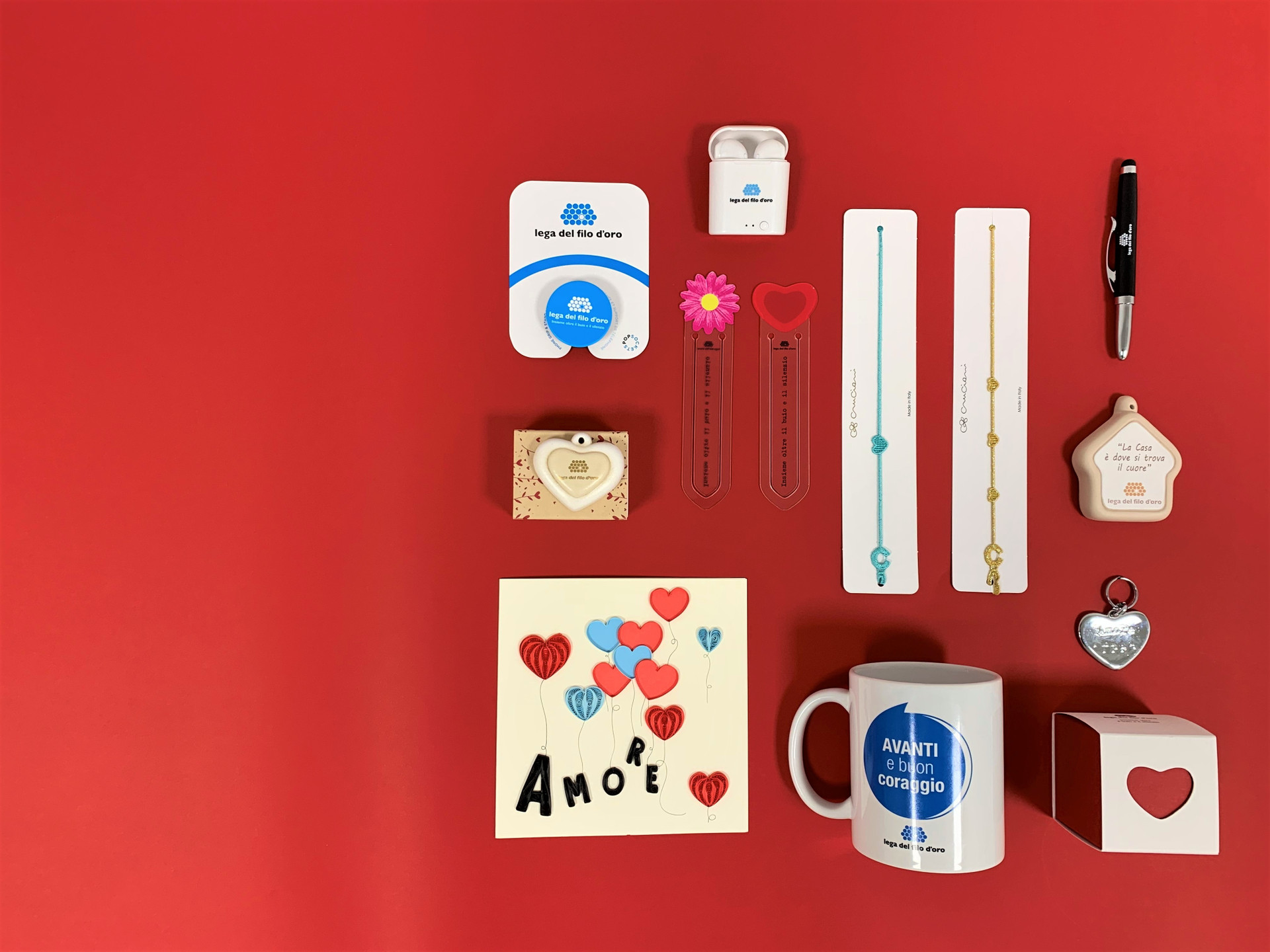 Composizione di regali solidali per San Valentino su sfondo rosso, inclusi portachiavi a forma di cuore, biglietto "Amore", bomboniera a forma di cuore, casetta in ceramica, pop grip e auricolari wireless. Perfetti per donare con amore e solidarietà.