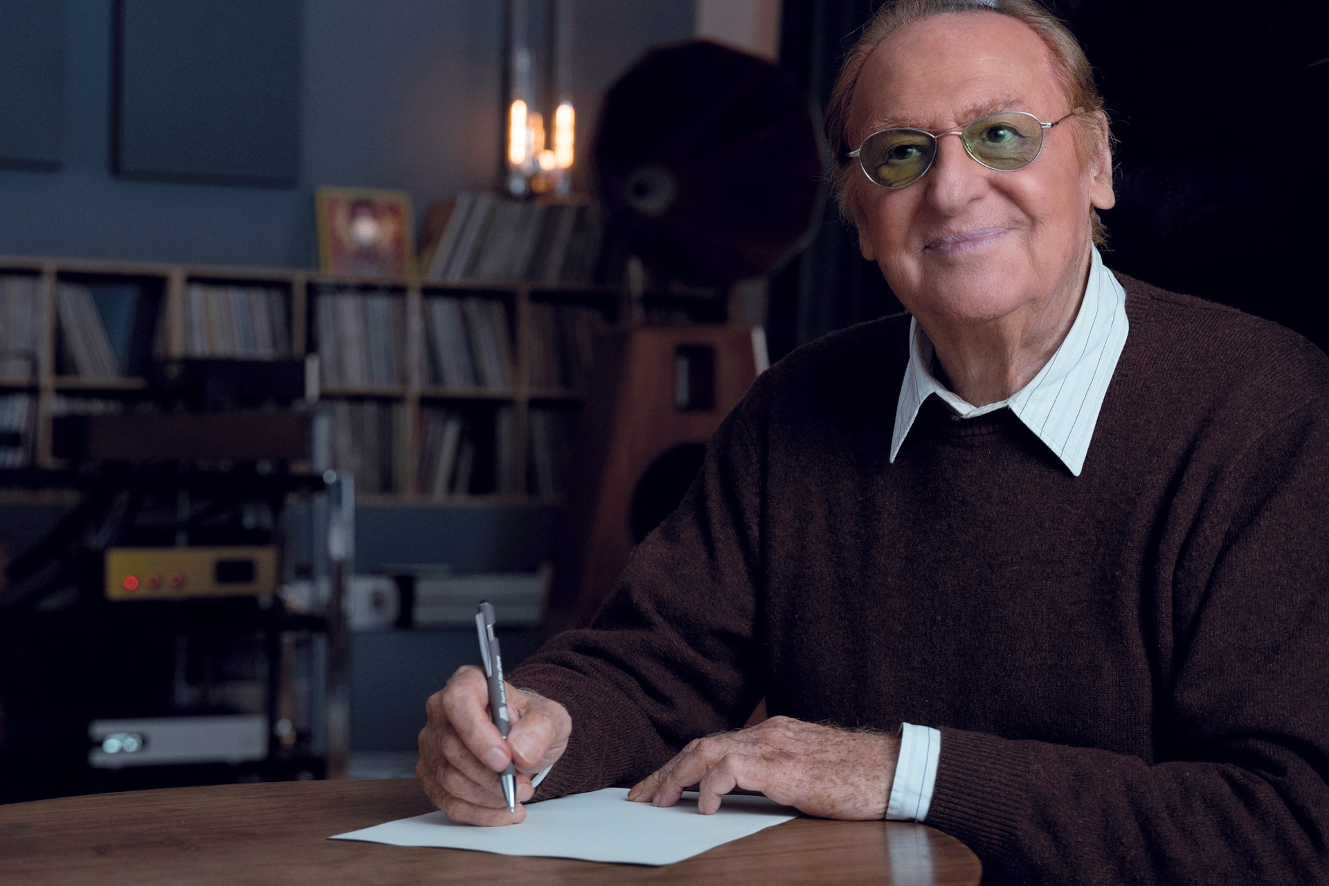 Renzo Arbore, testimonial della Fondazione Lega del Filo d'Oro posa sorridente, seduto alla scrivania mentre firma un foglio. 