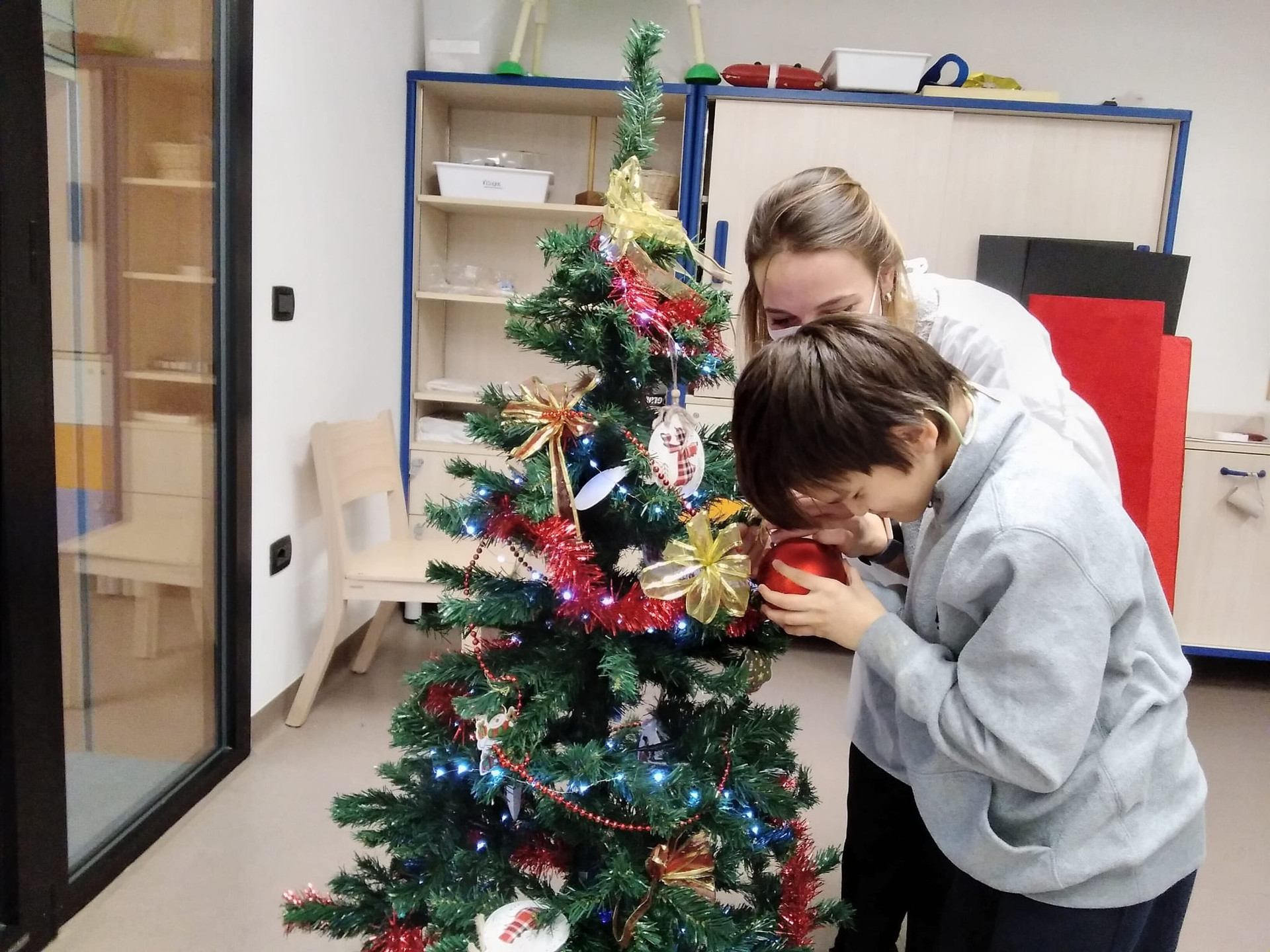 Un'educatrice aiuta un piccolo utente ad appendere una pallina rossa all'albero di Natale 