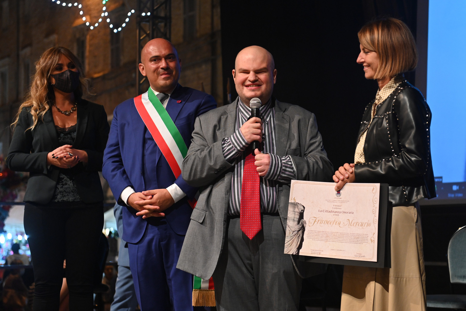 Francesco Mercurio, Presidente del Comitato delle Persone Sordocieche, è su un palco insieme ad altre tre persone: due donne e un uomo, che è il sindaco di Osimo. Il sindaco indossa la tipica fascia triocolore e tutti sono vestiti con abiti eleganti. Francesco Mercurio sorride e tiene in mano un microfono, mentre gli viene consegnata l'onorificenza.