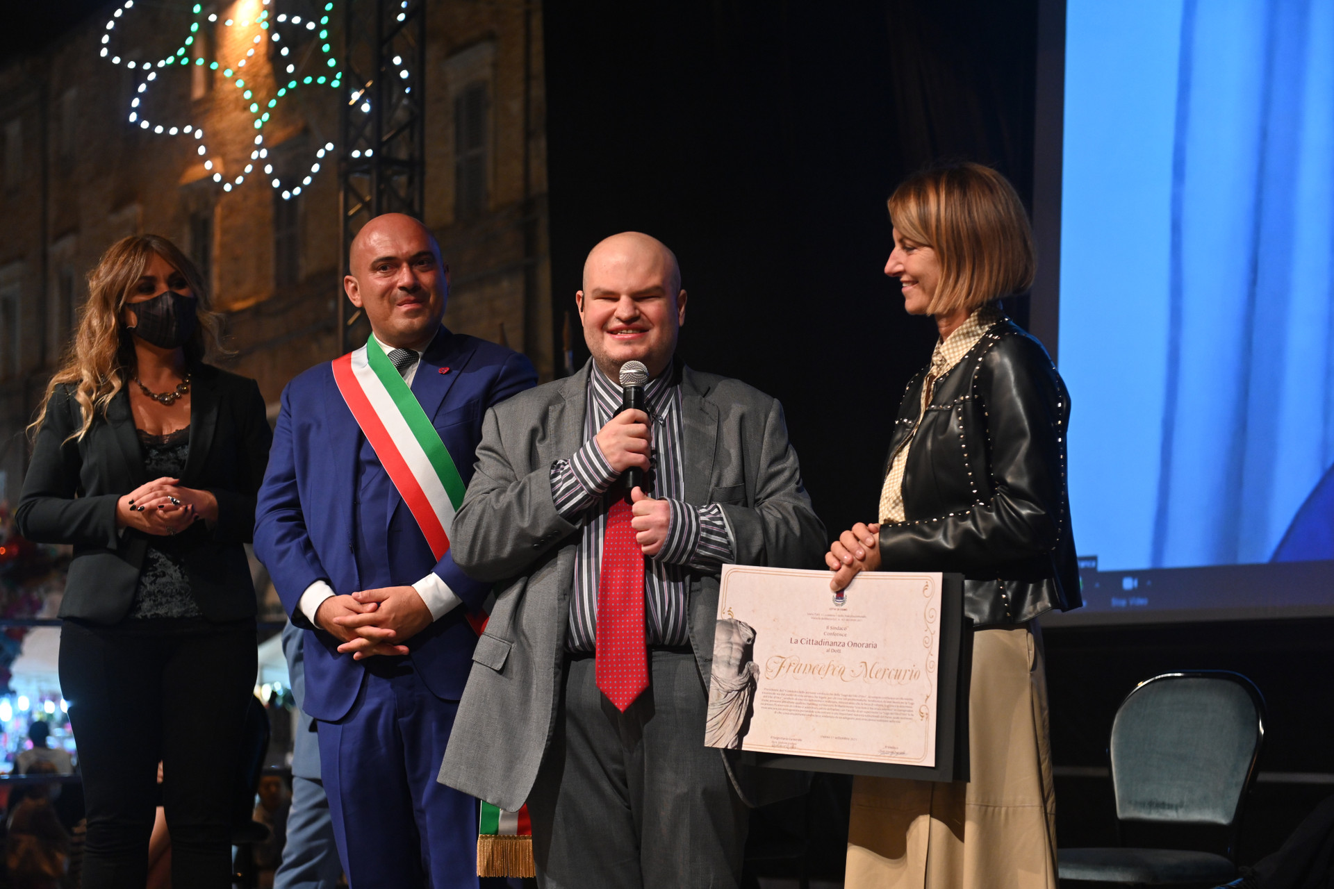 Francesco Mercurio, Presidente del Comitato delle Persone Sordocieche, è su un palco insieme ad altre tre persone: due donne e un uomo, che è il sindaco di Osimo. Il sindaco indossa la tipica fascia triocolore e tutti sono vestiti con abiti eleganti. Francesco Mercurio sorride e tiene in mano un microfono, mentre gli viene consegnata l'onorificenza.