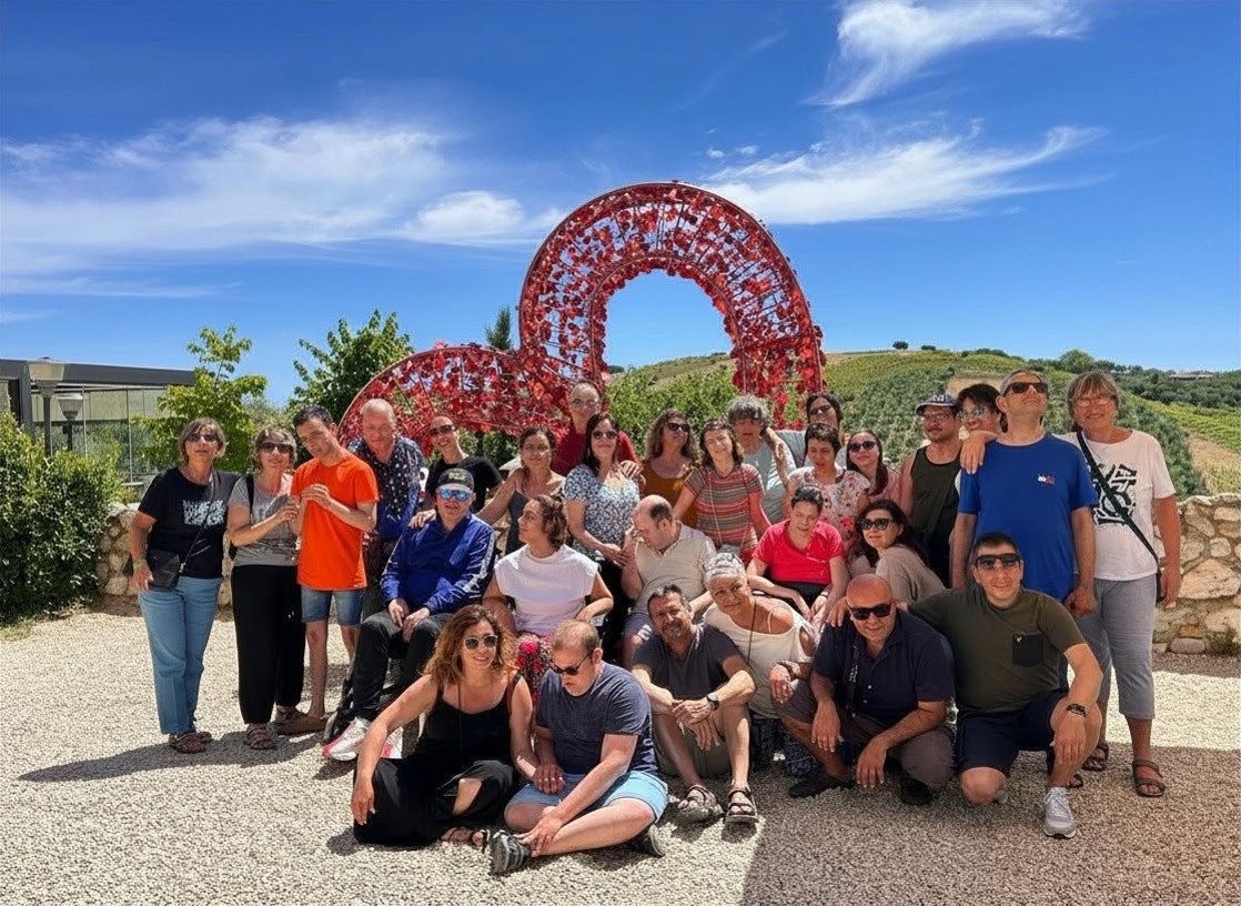 Foto di gruppo scattata in occasione del soggiorno estivo organizzato dalla Lega del Filo d'Oro a Giulianova, in aAbruzzo. Il gruppo di persone posa all’aperto su un piazzale ghiaioso. Alle spalle si trova una grande scultura rossa a forma di cuore intrecciato. Sullo sfondo si vedono colline verdi coltivate e un cielo azzurro con poche nuvole. 