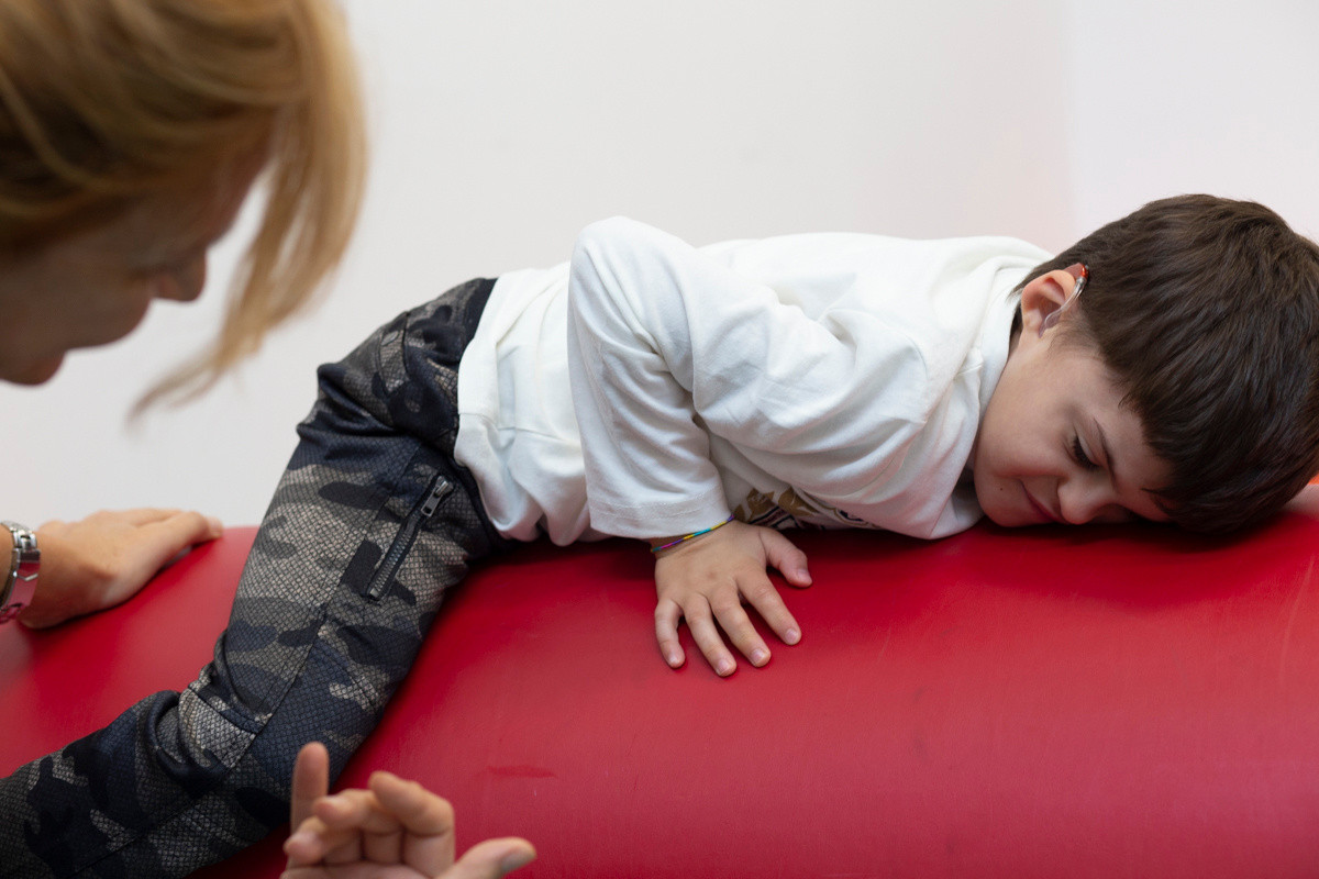 Agostino, bambino con sindrome di Charge seguito al Centro di Osimo, sta in equilibrio sdraiato su un cilindro di gommapiuma rosso, aiutato dalla fisioterapista Francesca