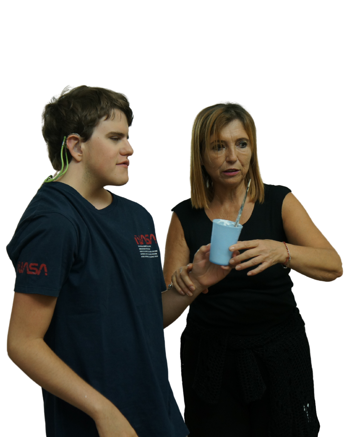 Foto senza sfondo. Thomas regge un bicchiere azzurro con dentro un pennello, al suo fianco l'educatrice lo guida mentre cammina .