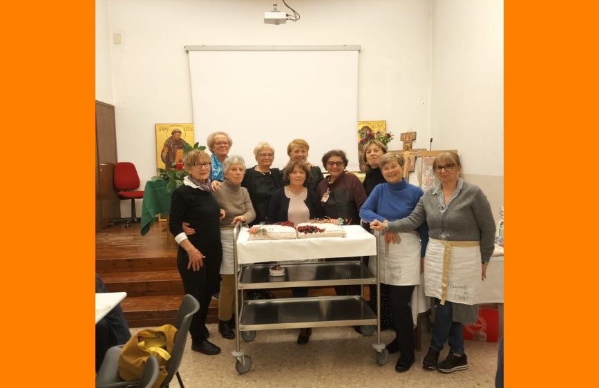 La foto ritrae un gruppo affiatato di donne, le “Amiche in cucina”, riunite attorno a un carrello con due torte decorate, pronte per essere condivise. Lo scatto trasmette un’atmosfera calda e conviviale, tipica di momenti trascorsi insieme tra ricette, sorrisi e passione per la cucina. Le donne, alcune con il grembiule indossato, sembrano legate da una forte amicizia e da un entusiasmo comune per l’arte culinaria, mentre l’ambiente semplice e familiare suggerisce un contesto di incontro e condivisione, forse all’interno di un'associazione o in occasione di un evento speciale.