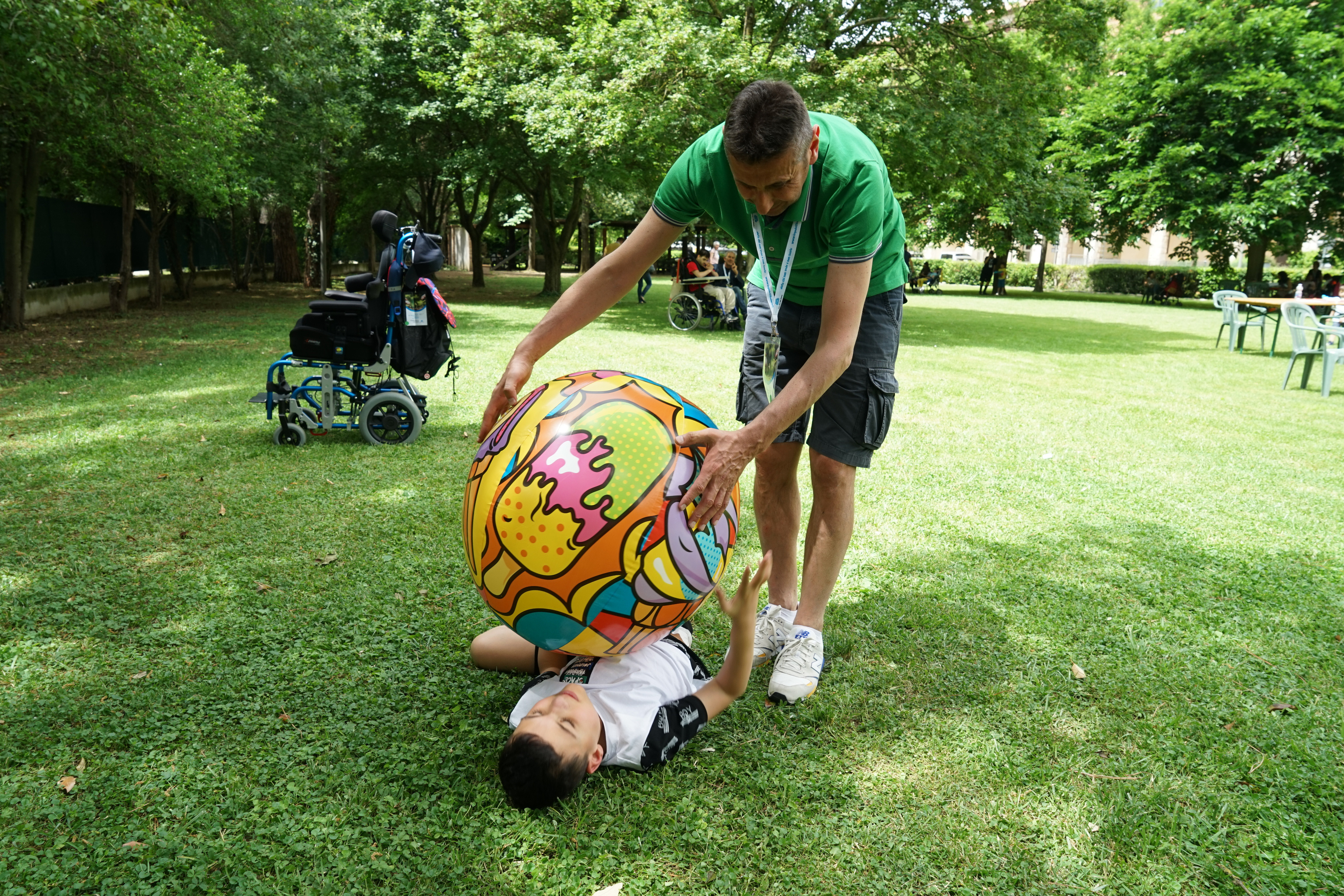 Un volontario e un piccolo utente giocano su un prato: il bambino è steso sull'erba mentre il ragazzo gli porge dall'alto una grande palla colorata