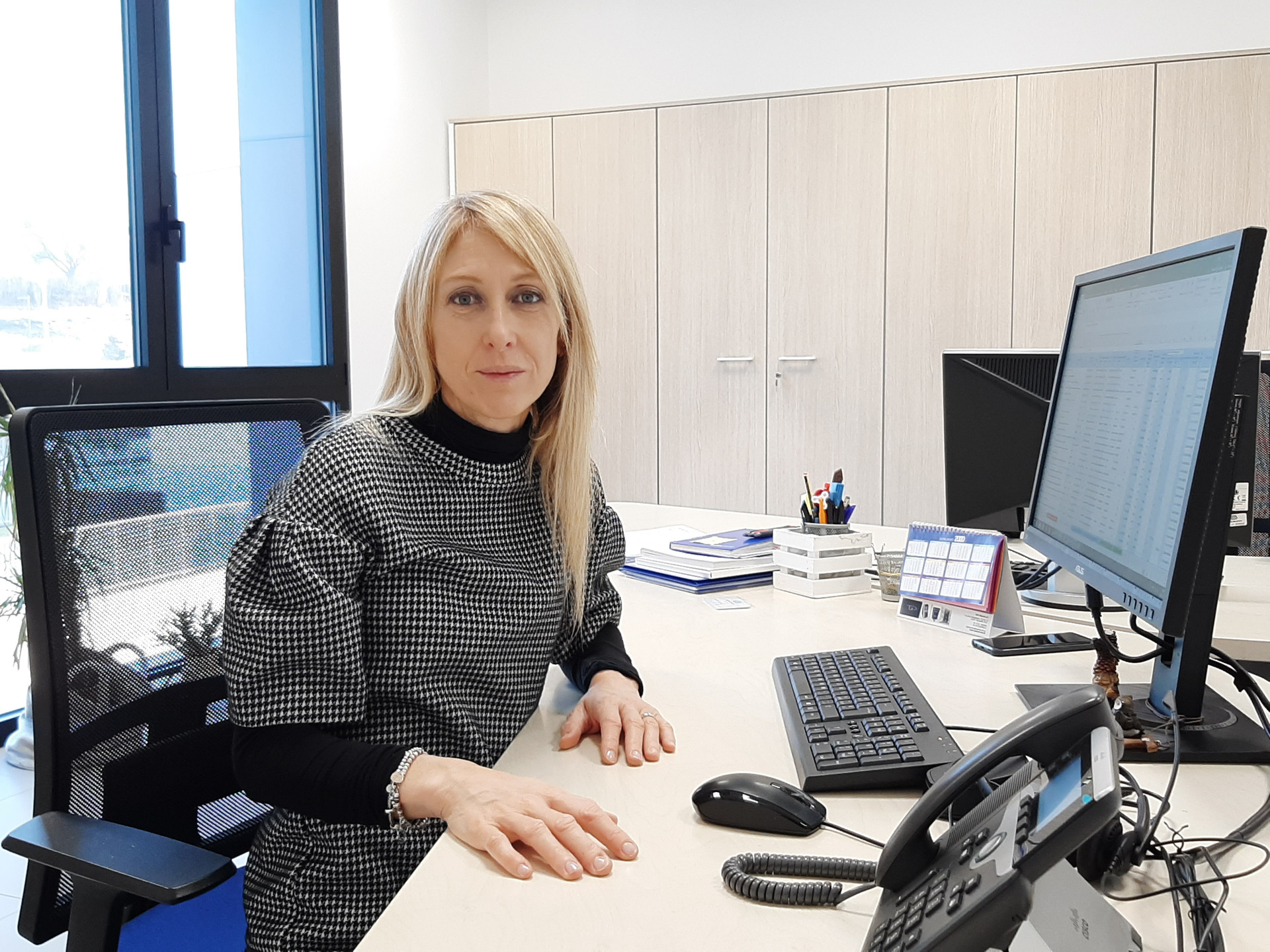 Simona è seduta alla sua scrivania. Davanti a lei ci sono tutti gli strumenti di lavoro: cancelleria, computer, monitor, tastiera, mouse e schermo.