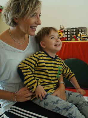 Edoardo, un bimbo utente della Lega del Filo d'Oro, è in braccio all'educatrice davanti ad uno specchio. Entrambi sorridono.