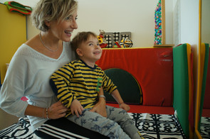 Edoardo, un bimbo utente della Lega del Filo d'Oro, è in braccio all'educatrice davanti ad uno specchio. Entrambi sorridono.