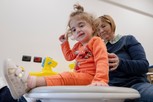 Benedetta, bambina con sindrome CHARGE seguita nel Centro di Osimo, esercita il suo senso dell'equilibrio seduta su una piattaforma basculante, con il sostegno della sua educatrice. Entrambe sorridono e Benedetta guarda chi la sta fotografando.