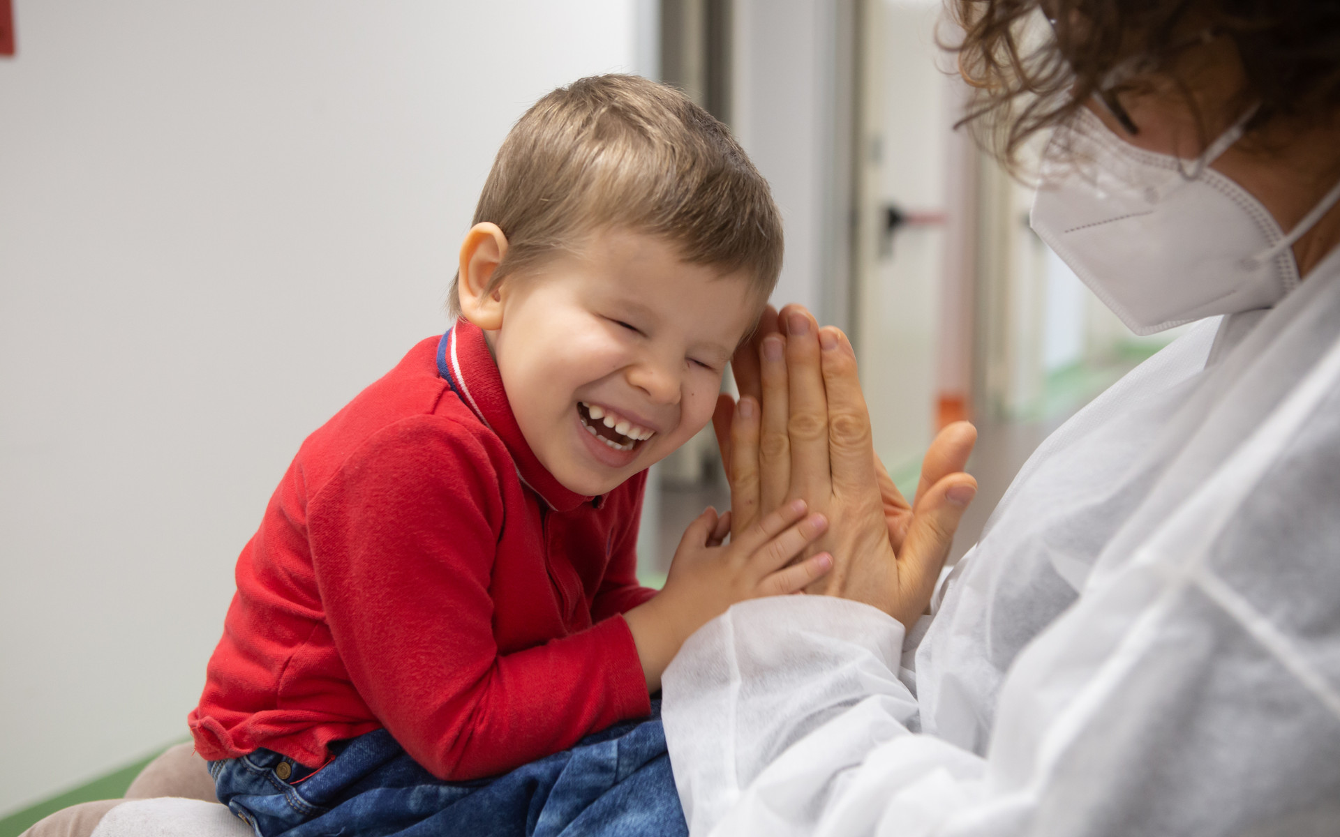 Matteo, piccolo utente del Centro Diagnostico, ride in braccio alla sua educatrice che batte le mani e gioca con lui. L'educatrice indossa camice monouso e mascherina chirurgica
