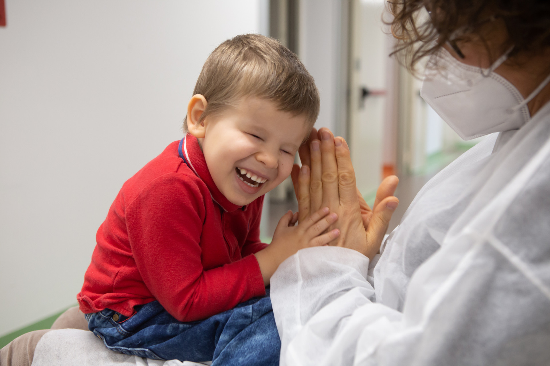 Matteo, piccolo utente del Centro Diagnostico, ride in braccio alla sua educatrice che batte le mani e gioca con lui. L'educatrice indossa camice monouso e mascherina chirurgica