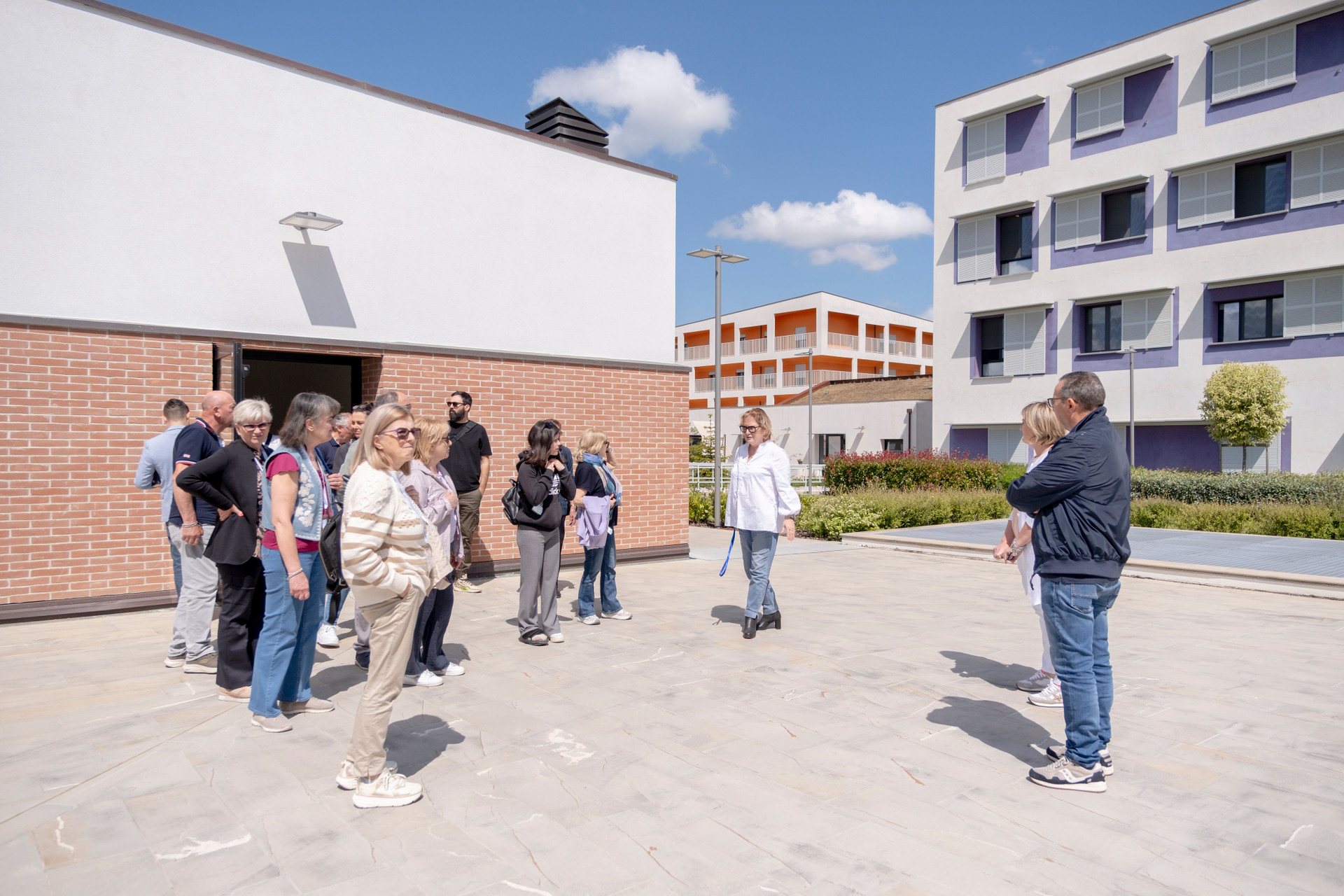 Un gruppo di persone visita gli spazi esterni del Centro Nazionale della Lega del Filo d’Oro in una giornata soleggiata. L’area è ampia, pavimentata e circondata da edifici moderni dai colori chiari e accesi (giallo, viola, arancione). Le persone, di varie età, ascoltano con interesse una guida mentre osservano l’ambiente circostante. Sullo sfondo, siepi curate e cielo azzurro con qualche nuvola soffice completano l’atmosfera accogliente e ordinata del luogo. 