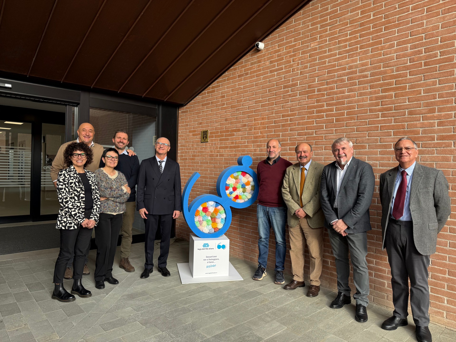 Foto di gruppo scatatta in oocasione della visita dell'Assessore alla Sanità della Regione Marche, Paolo Calcinaro, presso il Centro Nazionale della Lega del Filo d’Oro ETS. La foto, scatatta all'ingresso del centro nazionale accanto al totem celebrativo dei 60 anni, ritrae l'assessore, il Presidente, il Direttore Generale, il Consigliere Roberto Oreficini Rosi, e altri Dirigenti e Responsabili dell’Ente insieme ai rappresentanti dei familiari e delle persone sordocieche.