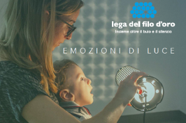 Il piccole Gabriele è in braccio a una donna, che regge una lampada accesa. La luce si riflette sul muro. La scritta "Emozioni di Luce" e il logo della Lega del Filo d'Oro sono nell'angolo in alto a destra.