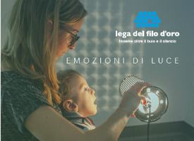 Il piccole Gabriele è in braccio a una donna, che regge una lampada accesa. La luce si riflette sul muro. La scritta "Emozioni di Luce" e il logo della Lega del Filo d'Oro sono nell'angolo in alto a destra.