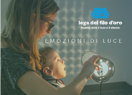Il piccole Gabriele è in braccio a una donna, che regge una lampada accesa. La luce si riflette sul muro. La scritta "Emozioni di Luce" e il logo della Lega del Filo d'Oro sono nell'angolo in alto a destra.