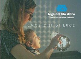 Il piccole Gabriele è in braccio a una donna, che regge una lampada accesa. La luce si riflette sul muro. La scritta "Emozioni di Luce" e il logo della Lega del Filo d'Oro sono nell'angolo in alto a destra.