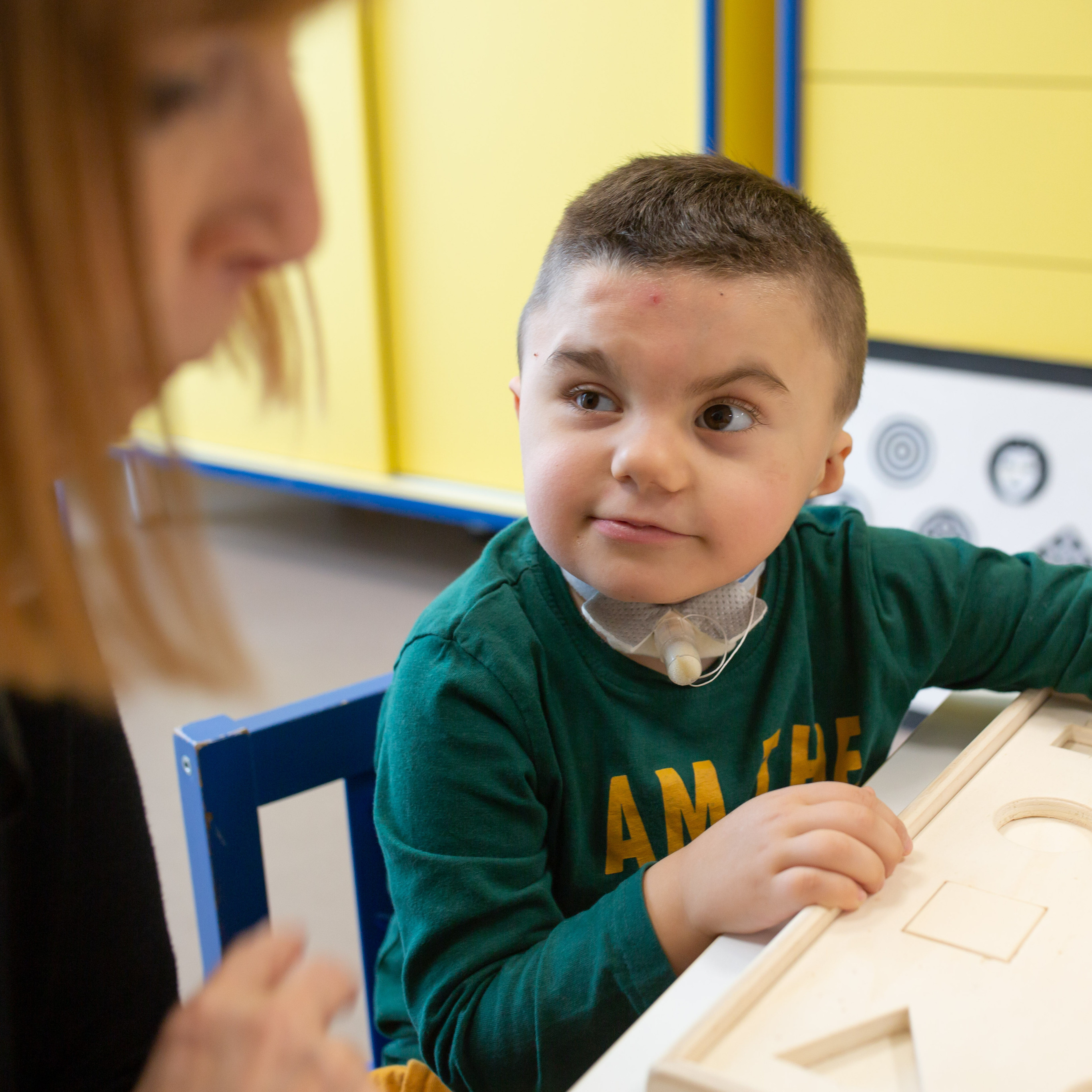 Il piccolo Leonardo, bambino con sindrome di Charge a trattamento nel Centro Nazionale, guarda su verso l'educatrice mentre fa un'attività con telle sagome in legno