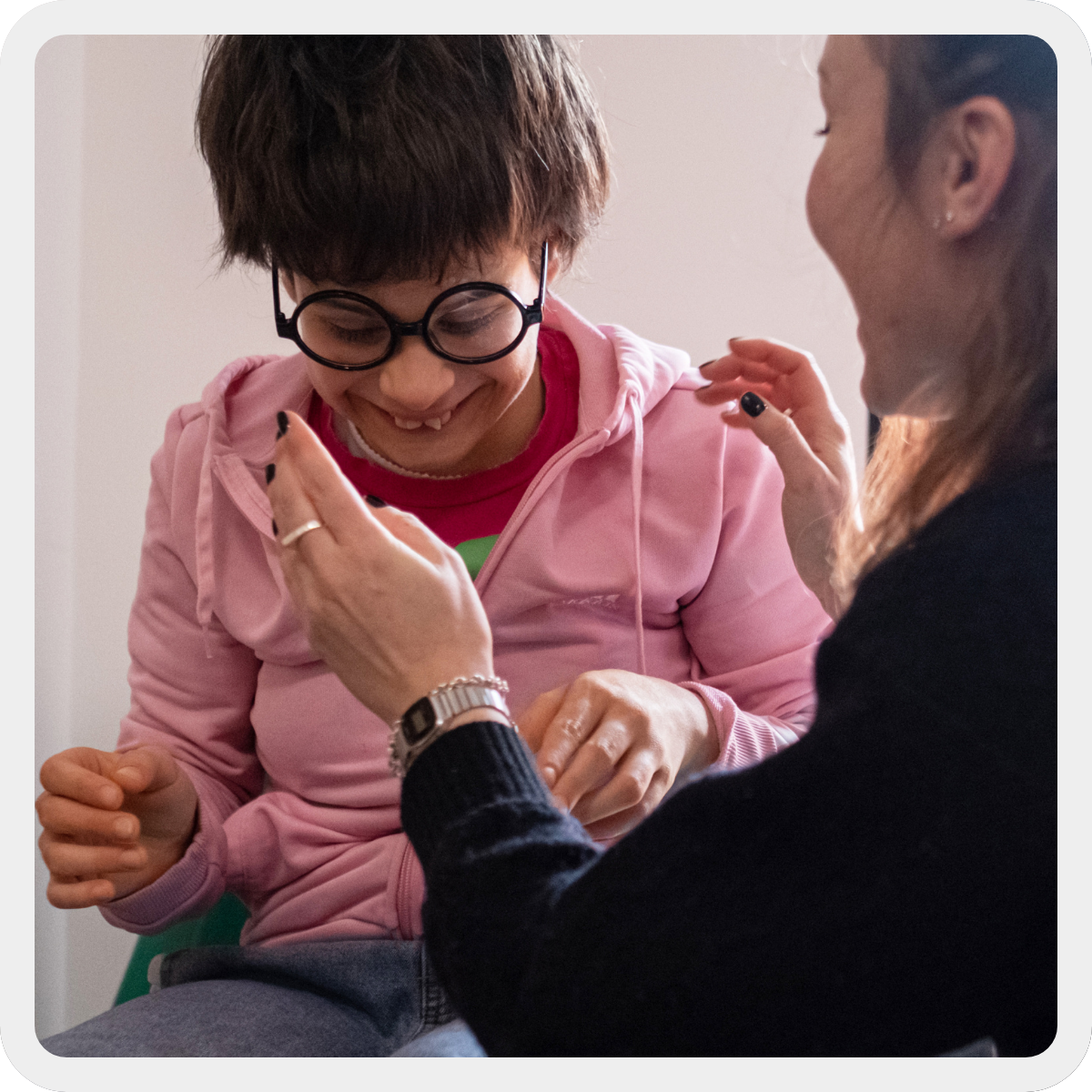 Nicole, ragazza con pluridisabuilità di circa 15 anni, gioca con la sua educatrice e sorride divertita. Indossa scherzando un paio di occhiali tondi e neri. 