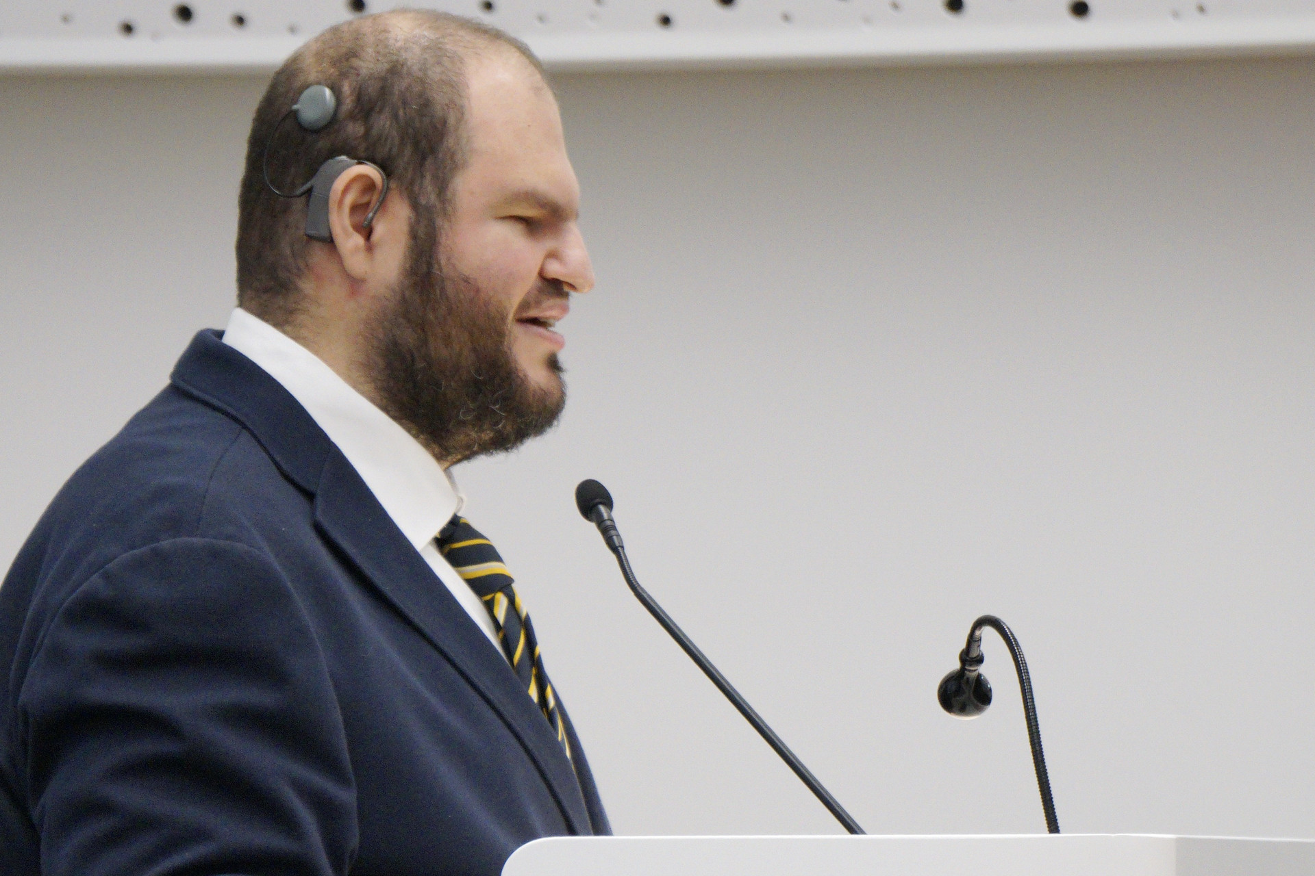 Francesco Mercurio, Presidente del Comitato delle Persone Sordocieche, è in piedi davanti ad un piedistallo con due microfoni. È vestito in modo formale.