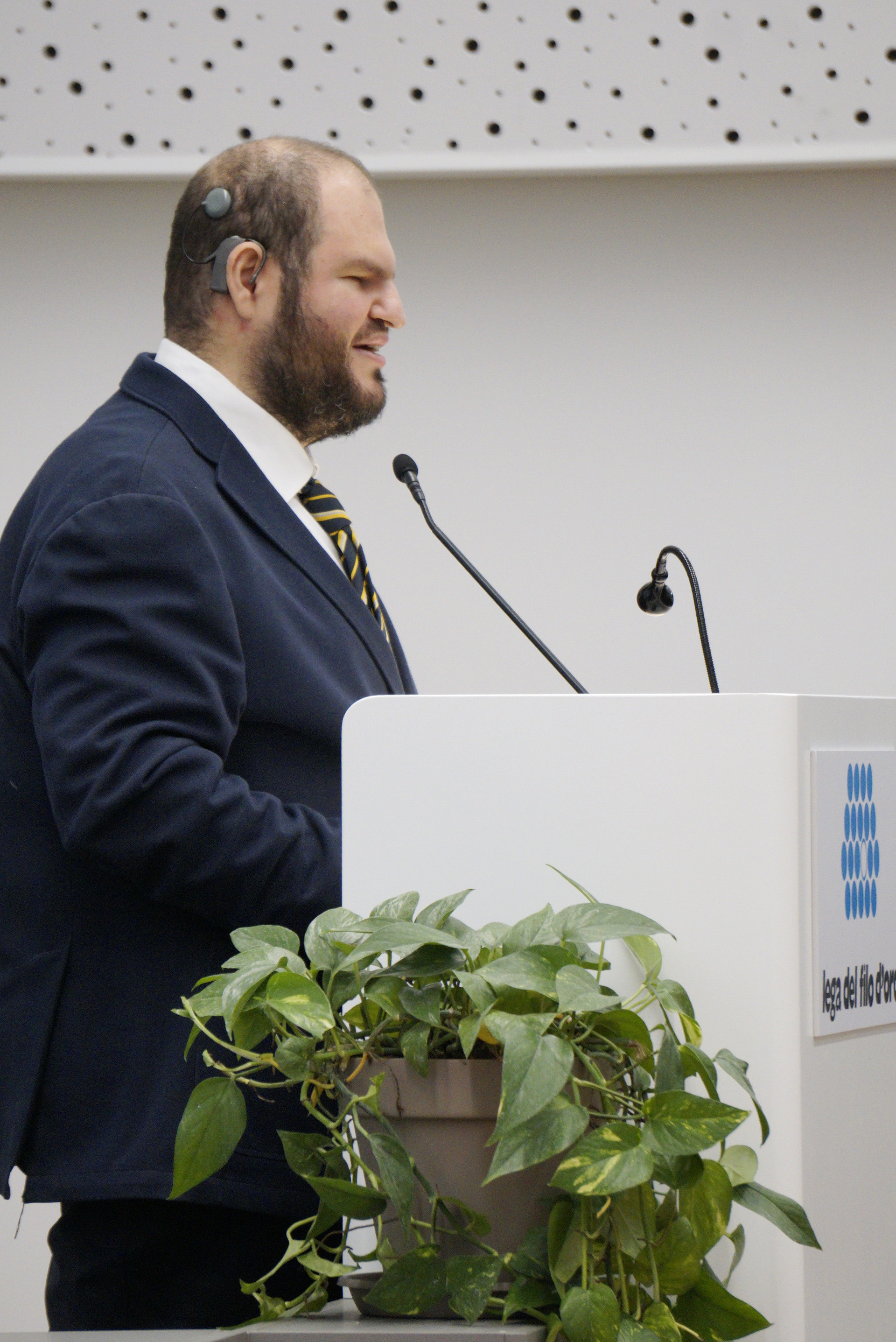 Francesco Mercurio, Presidente del Comitato delle Persone Sordocieche, è in piedi davanti ad un piedistallo con due microfoni. È vestito in modo formale. 