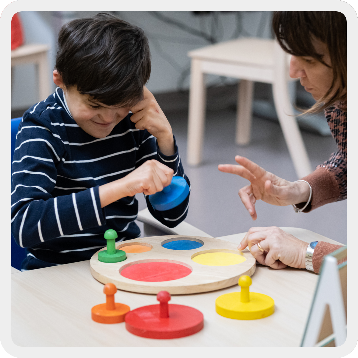 Agostino, bambino con la sindrome di Charge, fa attività di incastro con un gioco di legno colorato. La sua educatrice Lorenza gli fa il segno LIS "bravo" perchè sta mettendo il cerchio blu nel posto corretto. 