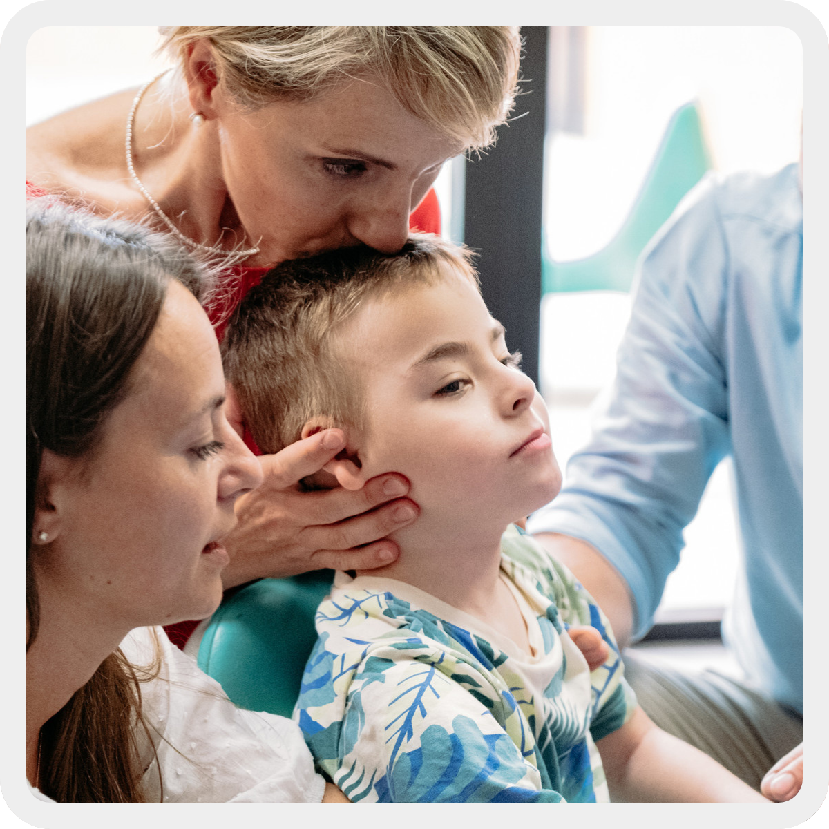 Edoardo, bambino di circa 10 anni con pluridisabilità, riceve un bacio sulla testa dalla sua educatrice Manola che lo sorregge da dietro. Alla sua destra c'è la sua mamma Caterina e alla sua sinistra si intravede il braccio di suo papà Giovanni. 