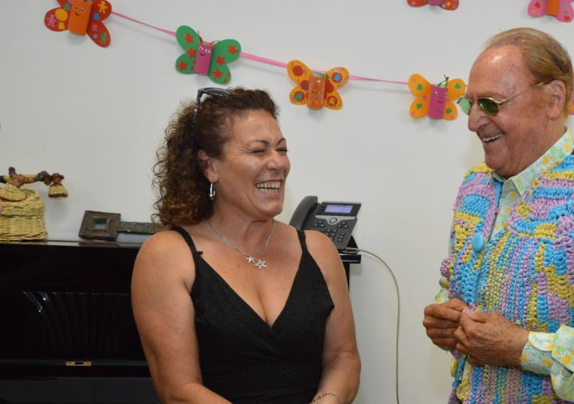 Renzo Arbore felice in visita alla sede di Napoli riceve in dono da Matilde, sorridente ed emozionata, un colorato gilet realizzato a mano da lei. Accanto ad Arbore è presente anche il Presidente Rossano Bartoli che sorride.