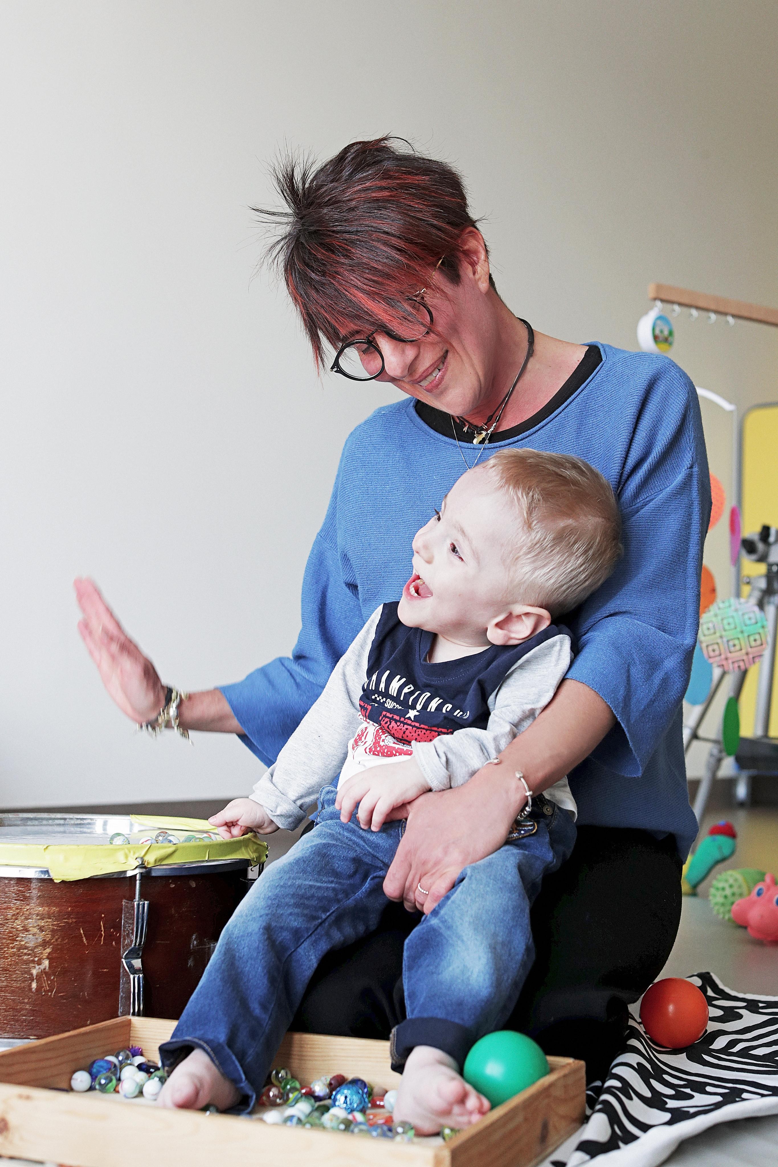 Il piccolo Gabriele ride di gusto in braccio alla sua educatrice che batte su un tamburo, mentre il bimbo muove i piedini in una scatola piena di biglie e palline colorate