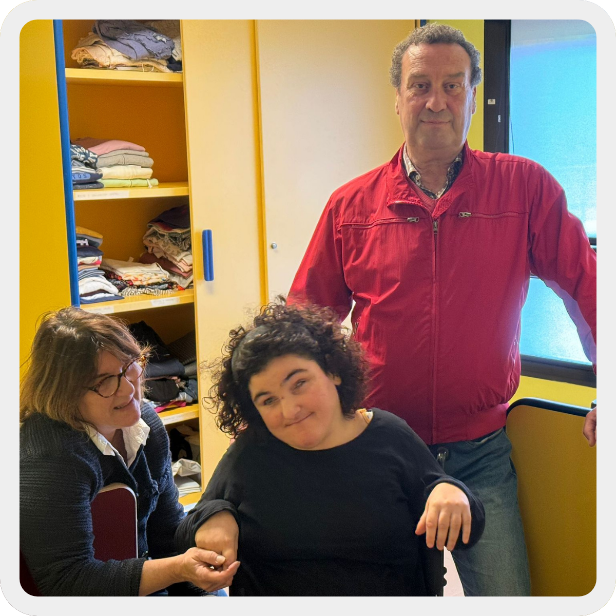 Sara, ragazza con pluridisabilità di circa 20 anni, è con i suoi genitori Anna e Daniele all'interno della sua stanza al Centro di Lesmo. Sullo Sfondo un armadio giallo con maniglie e bordi blu che contiene i suoi vestiti (che si intravedono) 
