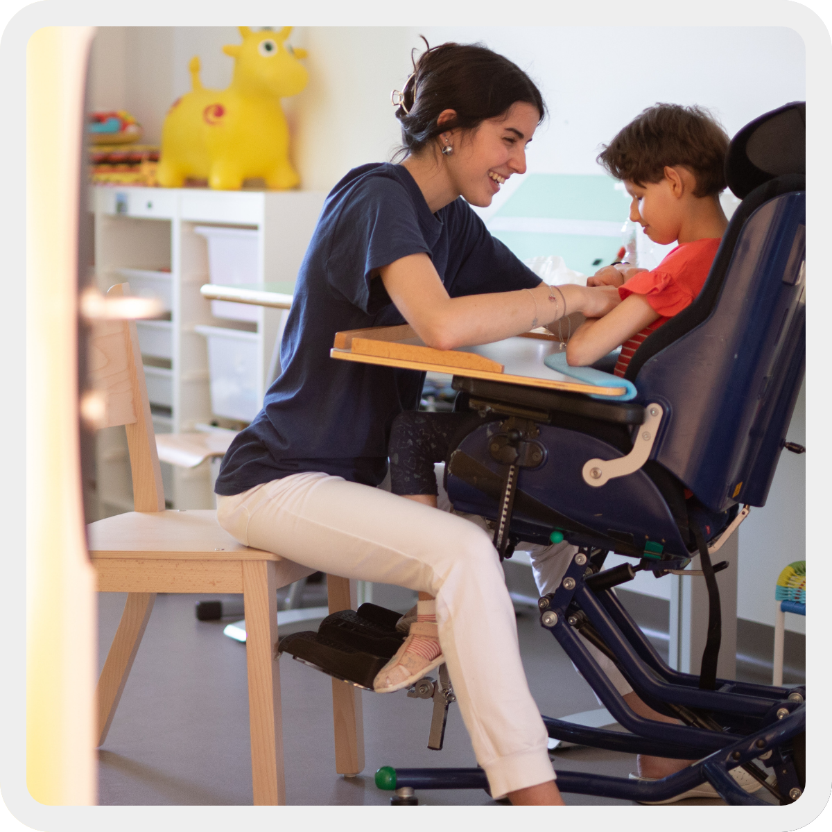 Asia, bambina con pluridisabilità, è seduta sul suo sistema postrurale. Di fronte a lei la sua logopedista che sorride mentre le tiene le mani. 