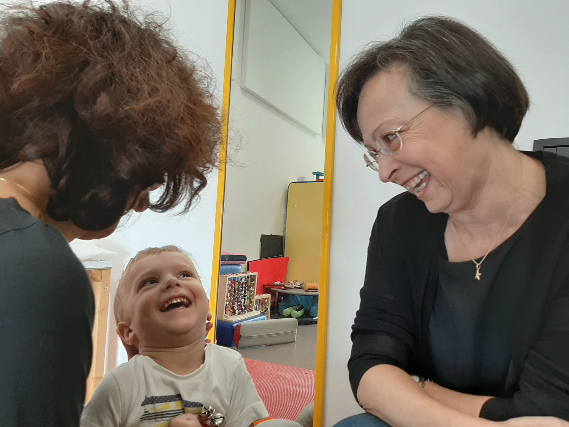 Un bambino utente della Lega del Filo d'Oro ride al suono dei sonagli insieme alla Dottoressa Raimondi e a Fiammetta la musicoterapista. La scena avviene in un ambiente dedicato alla musicoterapia