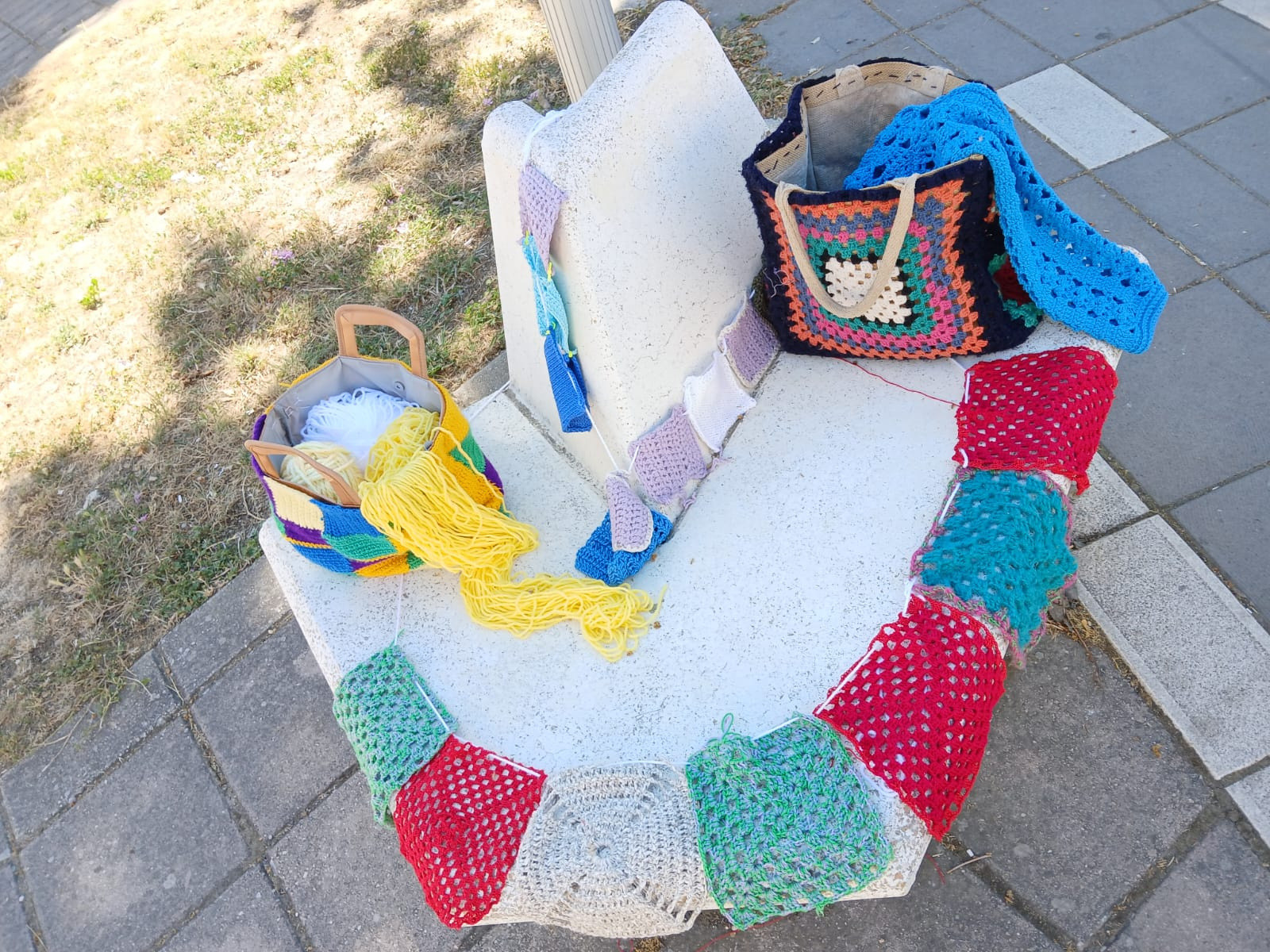 La Lega del Filo d’Oro ha aderito e promosso lo Yarn Bombing. Panchina decorata con creazioni colorate fatte a maglia. Sulla seduta sono appoggiate due borse, anch’esse realizzate a maglia