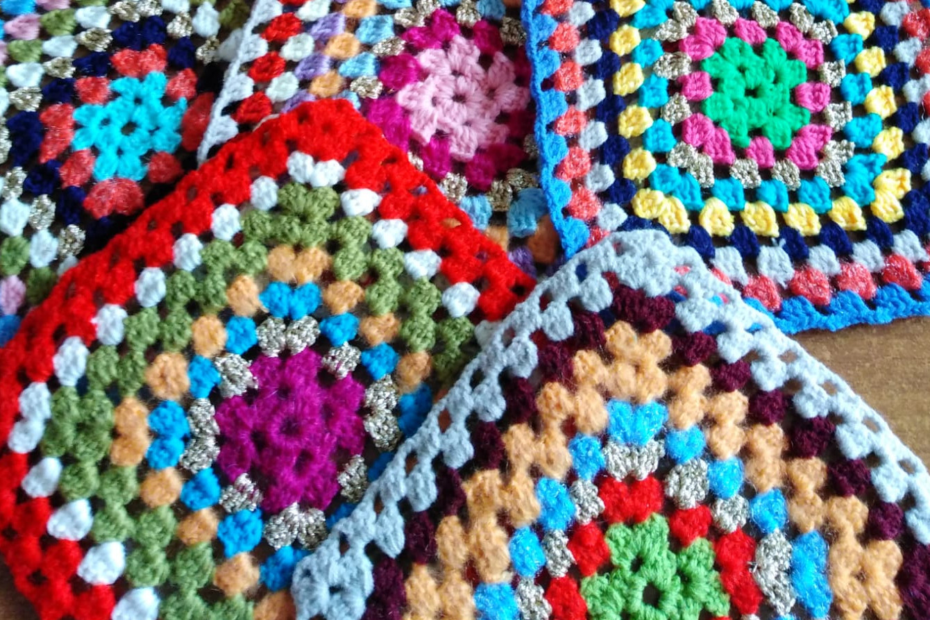 Dettaglio di mattonelle realizzate all’uncinetto con filati multicolore. Sullo sfondo due mani che lavorano all'uncinetto.