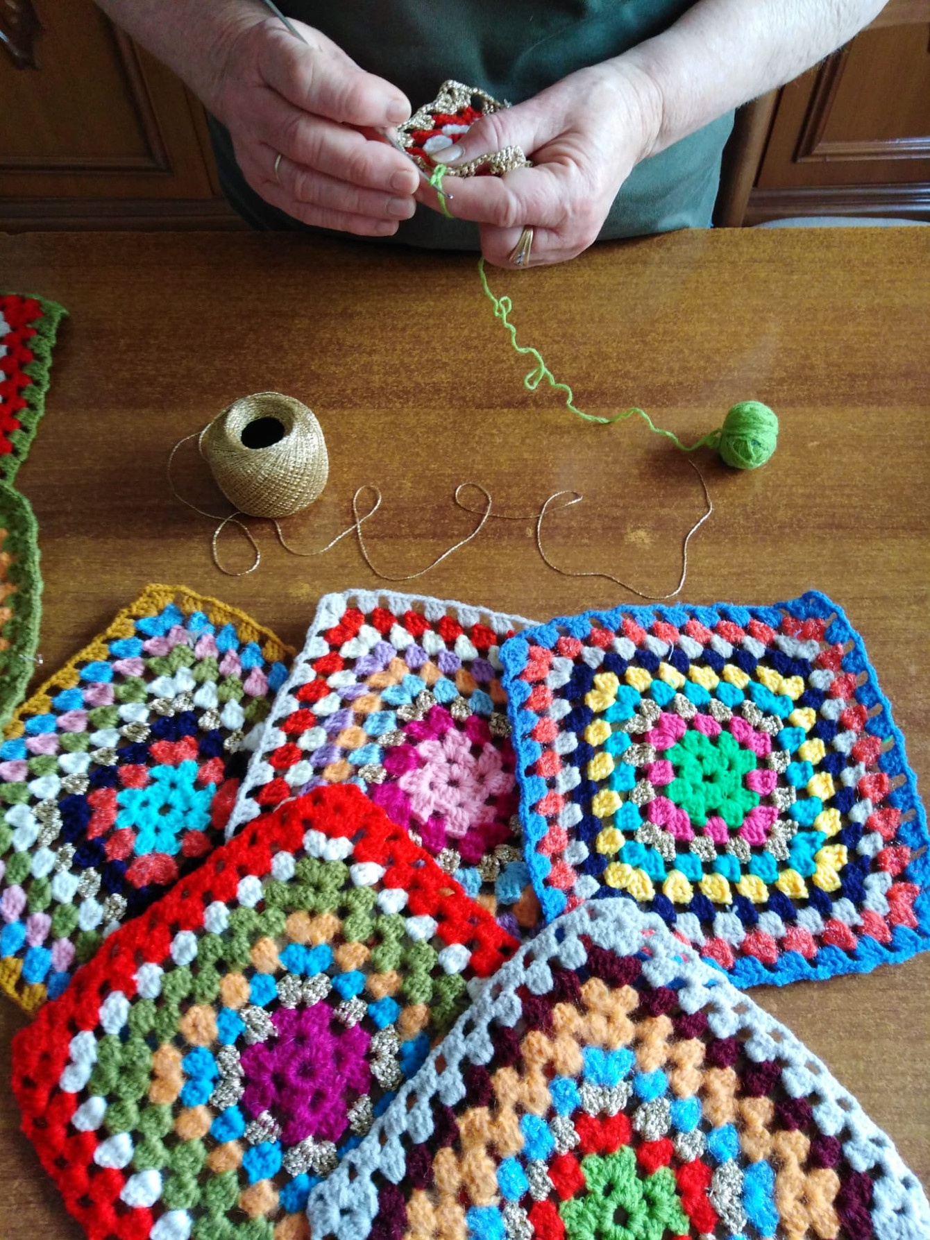 Dettaglio di mattonelle realizzate all’uncinetto con filati multicolore. Sullo sfondo due mani che lavorano all'uncinetto.