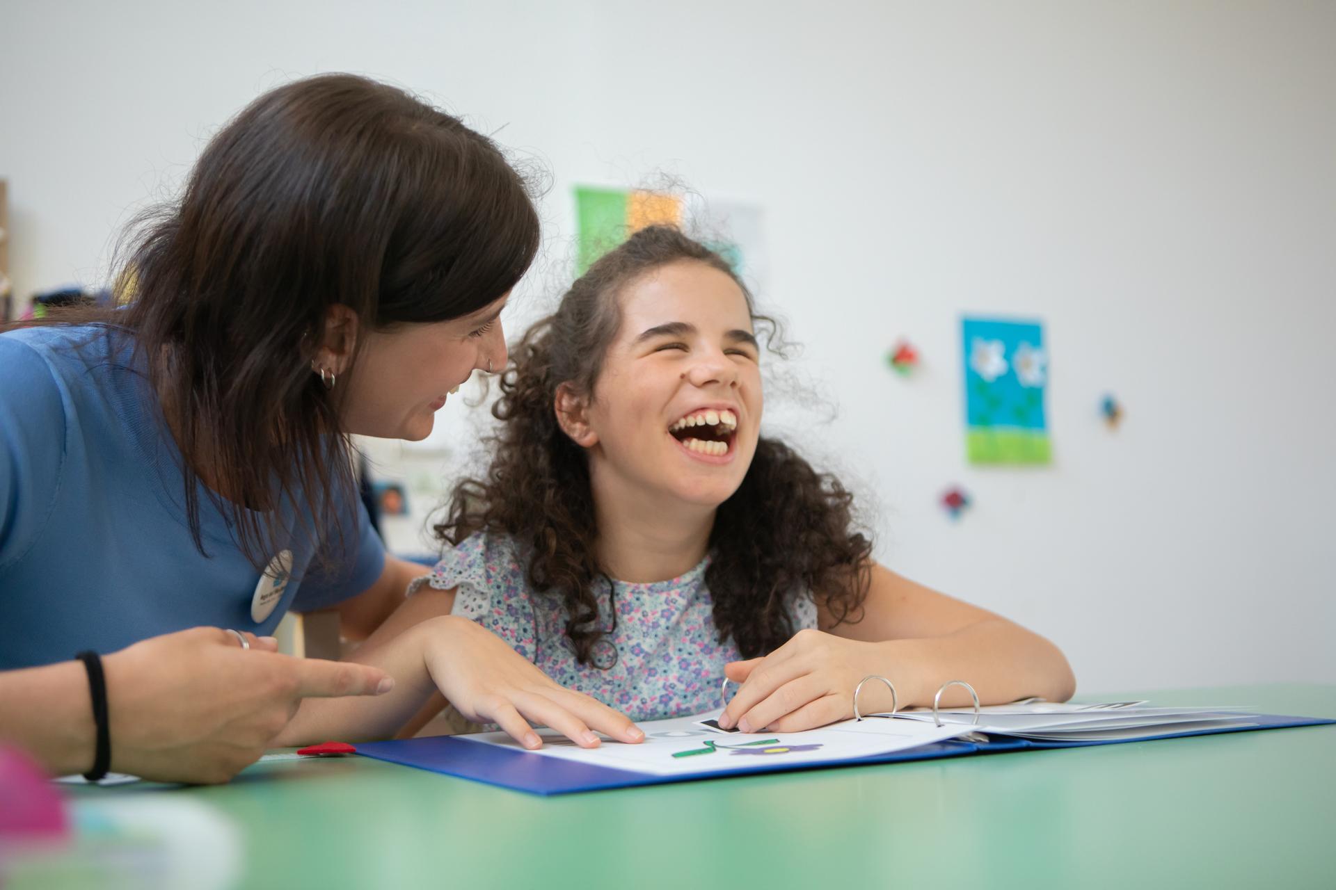 Giulia, una giovane ospite della Lega del Filo d’Oro, sorride felice accanto a un’operatrice. Entrambe sorridono davanti a una scrivania, mentre Giulia sfoglia un raccoglitore.