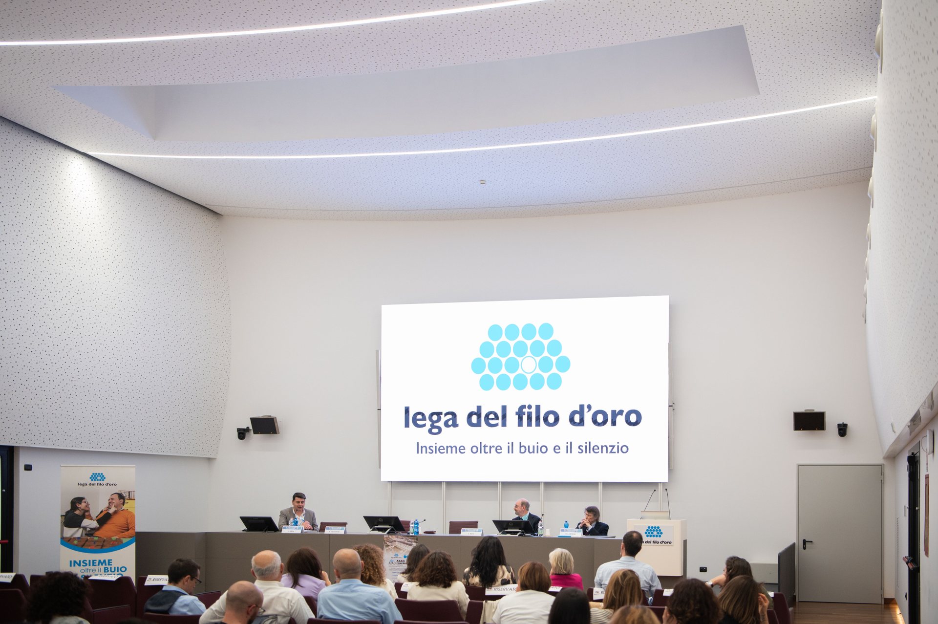 Terza Edizione della Conferenza Internazionale su Tecnologie Assistive e Disabilità. Sul maxischermo dell'auditorium compare il logo della Lega del Filo d'Oro.