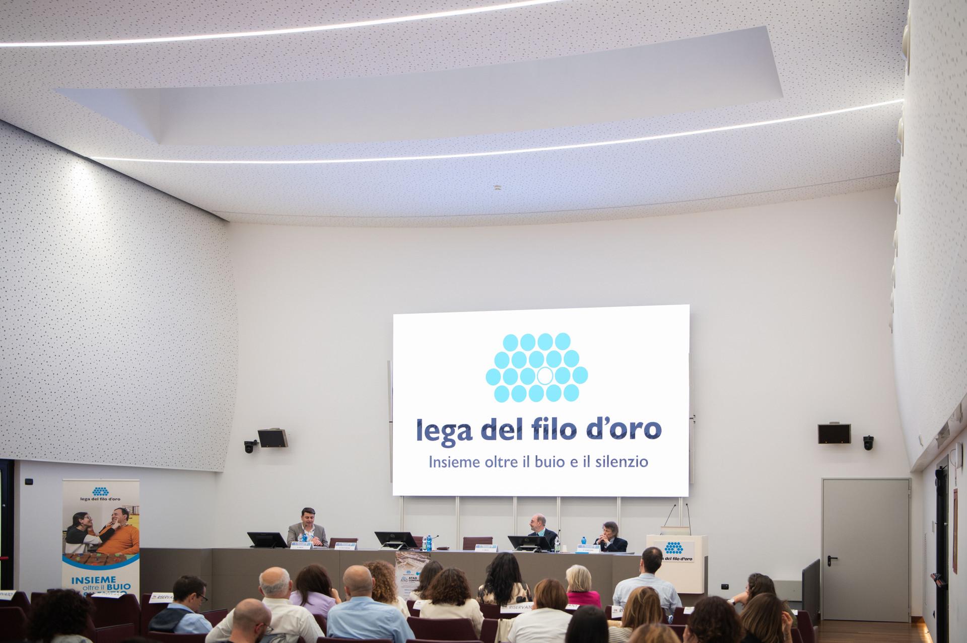 Terza Edizione della Conferenza Internazionale su Tecnologie Assistive e Disabilità. Sul maxischermo dell'auditorium compare il logo della Lega del Filo d'Oro.