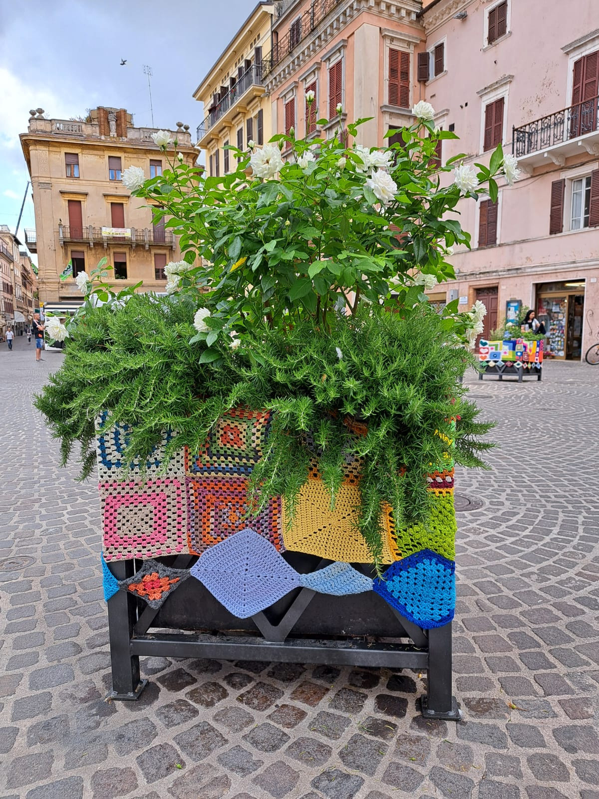 Grande vaso situato nella piazza di Osimo, decorato con vivaci mattonelle colorate fatte all’uncinetto. L’opera, parte dello Yarn Bombing promosso dalla Lega del Filo d’Oro.