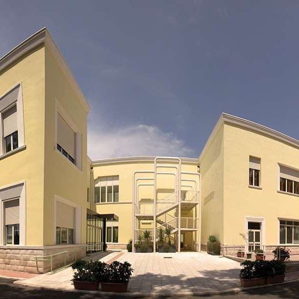 In grandangolo è fotografata il Centro Residenziale di Molfetta. Si vede in primo piano la strada, con una macchina che si allontana. Poi l'entrata piastrellata e tutto l'edificio, con i muri giallo limone. Il cielo è azzurro con qualche nuvola.