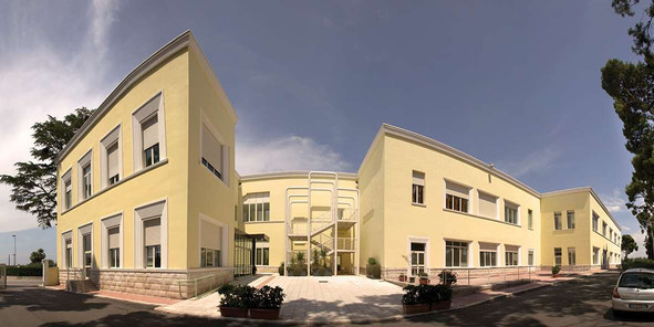 In grandangolo è fotografata il Centro Residenziale di Molfetta. Si vede in primo piano la strada, con una macchina che si allontana. Poi l'entrata piastrellata e tutto l'edificio, con i muri giallo limone. Il cielo è azzurro con qualche nuvola.