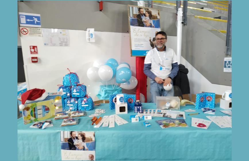 La foto mostra un uomo con la barba e gli occhiali che sorride, in piedi dietro un tavolo espositivo ricco di gadget e materiali promozionali, tra cui tazze, sacchetti regalo e palloncini bianchi e azzurri. Sullo sfondo si notano cartelli informativi e poster, suggerendo un evento di beneficenza o una fiera