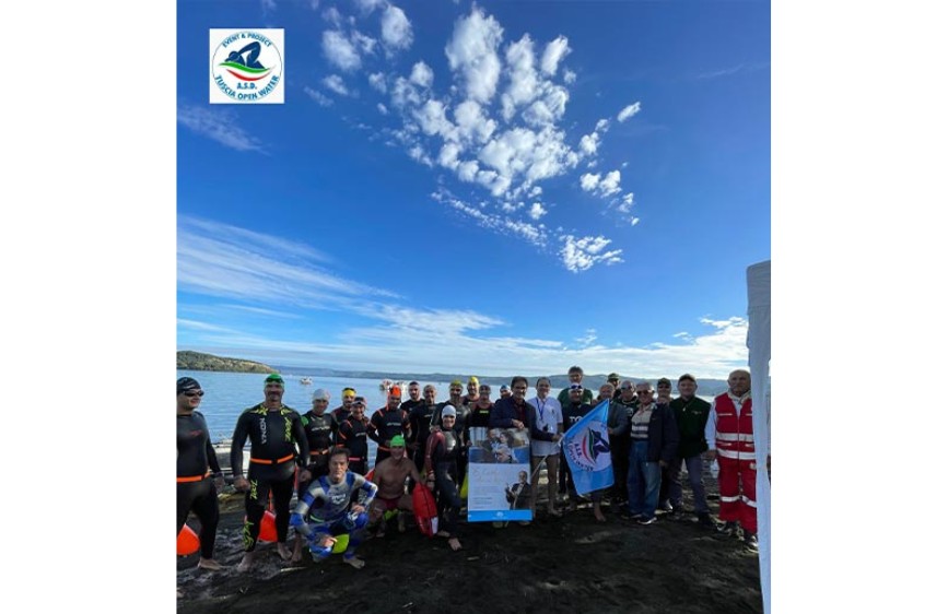 Foto di gruppo sulla spiaggia con partecipanti in abbigliamento da nuoto della Martana Open Water. Al centro viene esposta una locandina della Lega del Filo d’Oro. In alto a sinistra è visibile il logo dell’evento. Sullo sfondo, il lago e un cielo azzurro con nuvole.