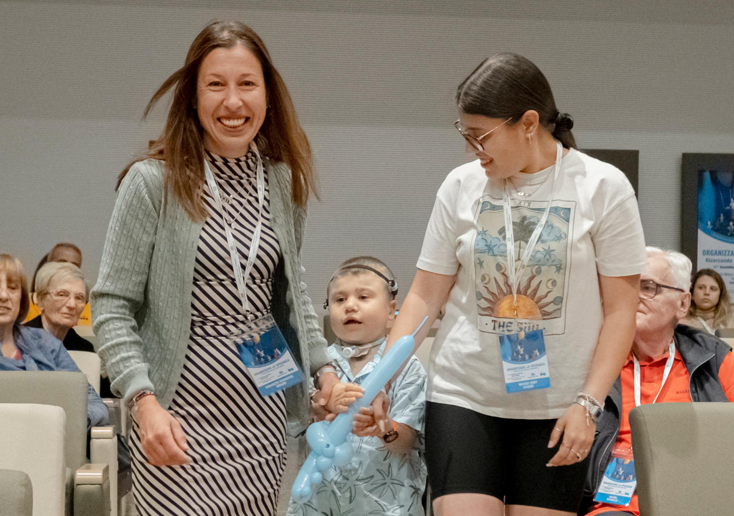 Mamma Shengyul, insieme al piccolo Berkay - bimbo seguito dalla Lega del FIlo d'Oro,e la sorella, camminano verso il palco durante l'Assemblea delle Famiglie. Ci sono tante persone sedute nella sala che sorridono rivolte verso di loro.