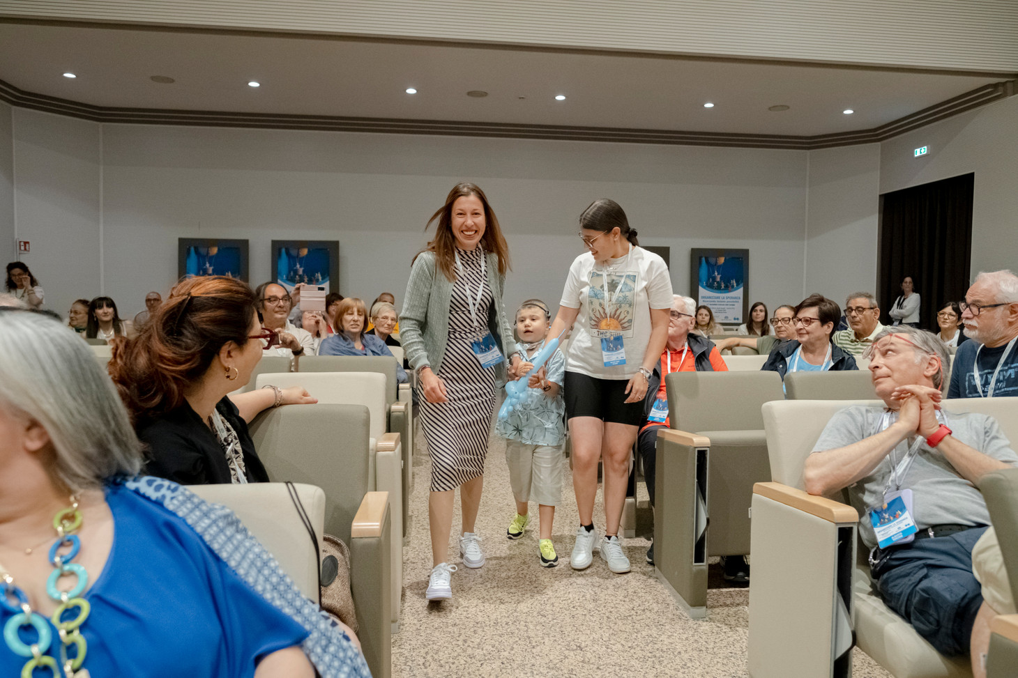 Mamma Shengyul, insieme al piccolo Berkay - bimbo seguito dalla Lega del FIlo d'Oro,e la sorella, camminano verso il palco durante l'Assemblea delle Famiglie. Ci sono tante persone sedute nella sala che sorridono rivolte verso di loro.