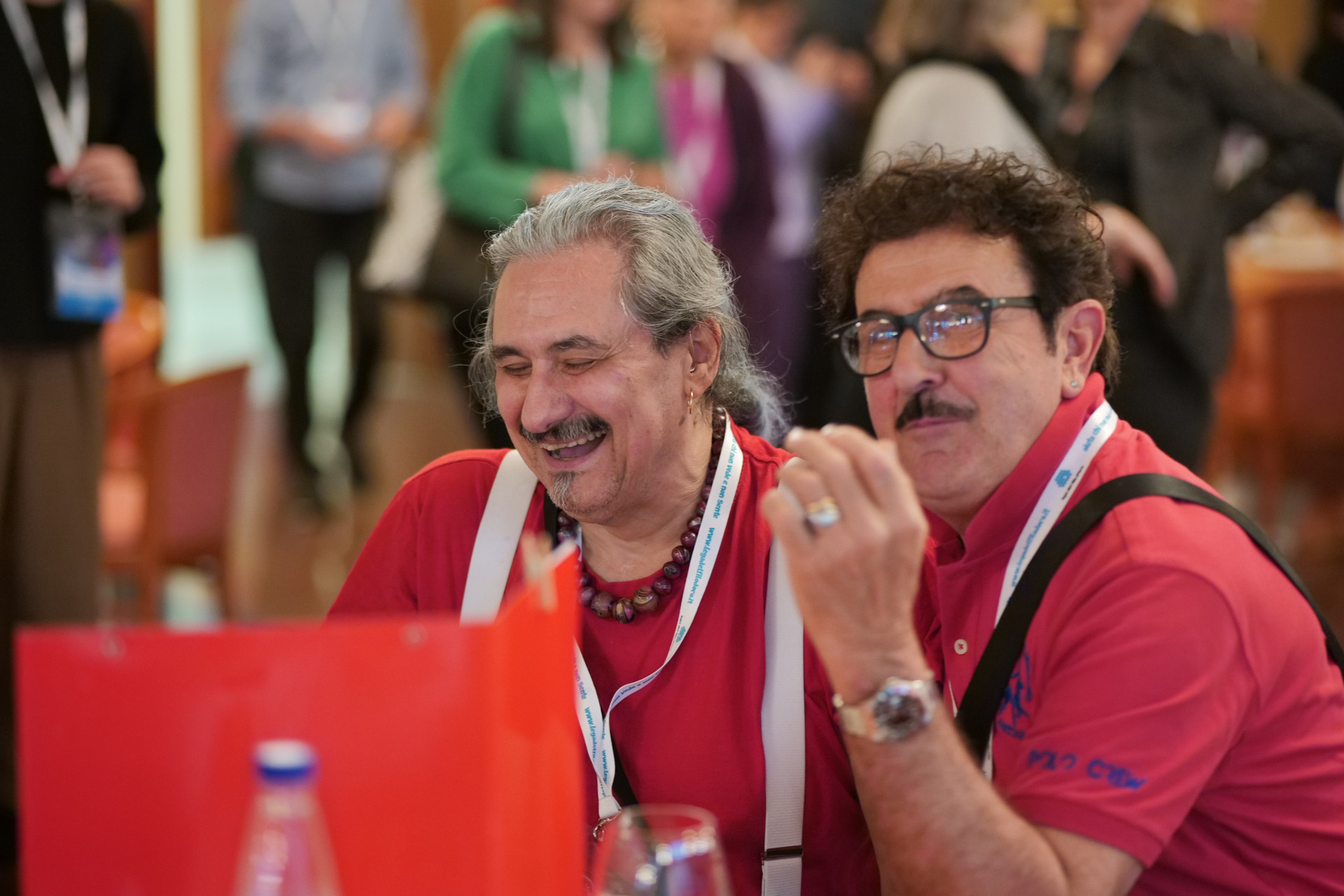 Roberto alla Conferenza delle Persone Sordocieche organizzata dalla Lega del Filo d'Oro a Tivoli nel 2023. La foto ritrae Roberto sorridente, al suo fianco un volontario guarda verso l'obiettivo. Sullo sfondo si vedono altre persone partecipanti alla Conferenza.