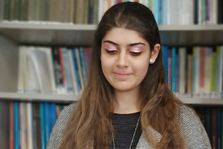 Lucia è fotografata all'interno di una biblioteca e si vedono gli scaffali pieni di libri dietro di lei. In questa foto è inquadrata a mezzo busto. È vestita in modo formale. Sta sorridendo.